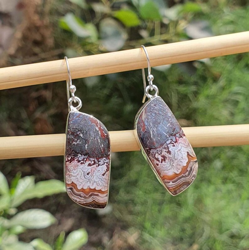 May include: A pair of dangle earrings featuring asymmetrical, polished stones with intricate, wavy banding in shades of dark brown, cream, and rust orange. The stones are set in silver-toned metal frames and hang from simple silver-toned shepherd hook wires.