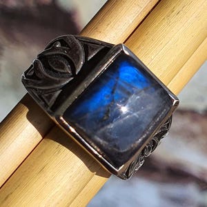 Può includere: Un anello d'argento con una pietra preziosa blu quadrata e iridescente. L'anello presenta dettagli ornati grigio scuro sui lati e intorno alla pietra. La pietra preziosa riflette la luce, mostrando una gamma di tonalità blu.