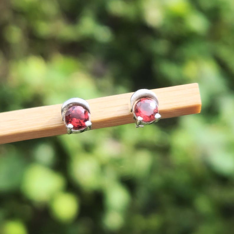 May include: Two small stud earrings with a silver setting, each holding a bright red gemstone. The earrings are presented on a light brown surface, set against a blurred green backdrop.
