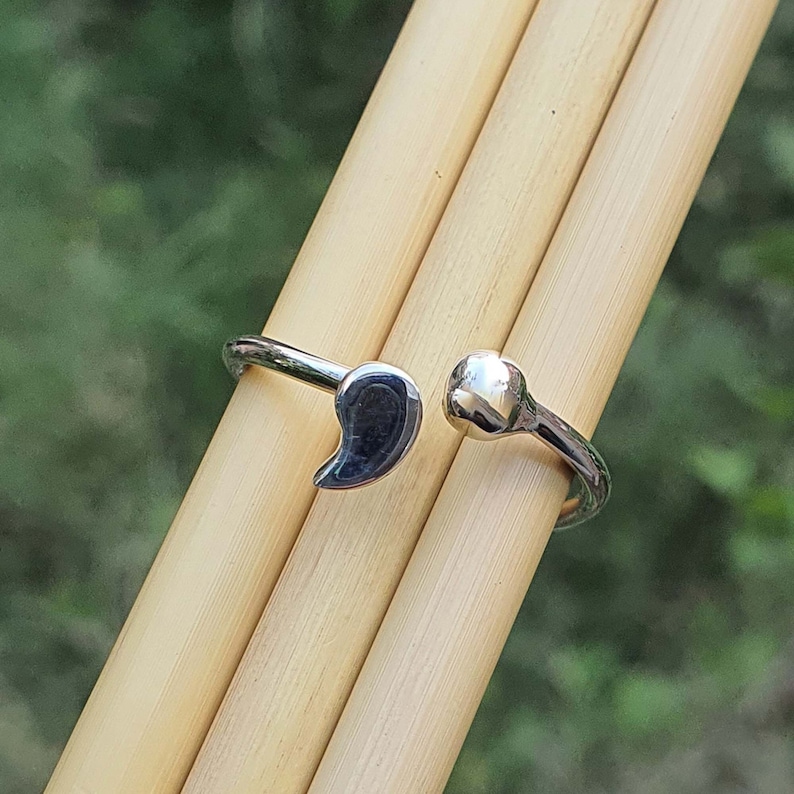 May include: A silver-toned open ring featuring a comma-shaped charm on one end and a small, smooth spherical bead on the other. The band is polished and metallic, designed with an adjustable gap between the two distinct terminal ends.
