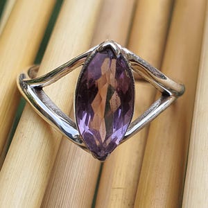 May include: A silver ring featuring a marquise-cut amethyst gemstone. The purple gemstone is set in a polished silver band with a unique, open design. The ring is displayed on a light brown surface.