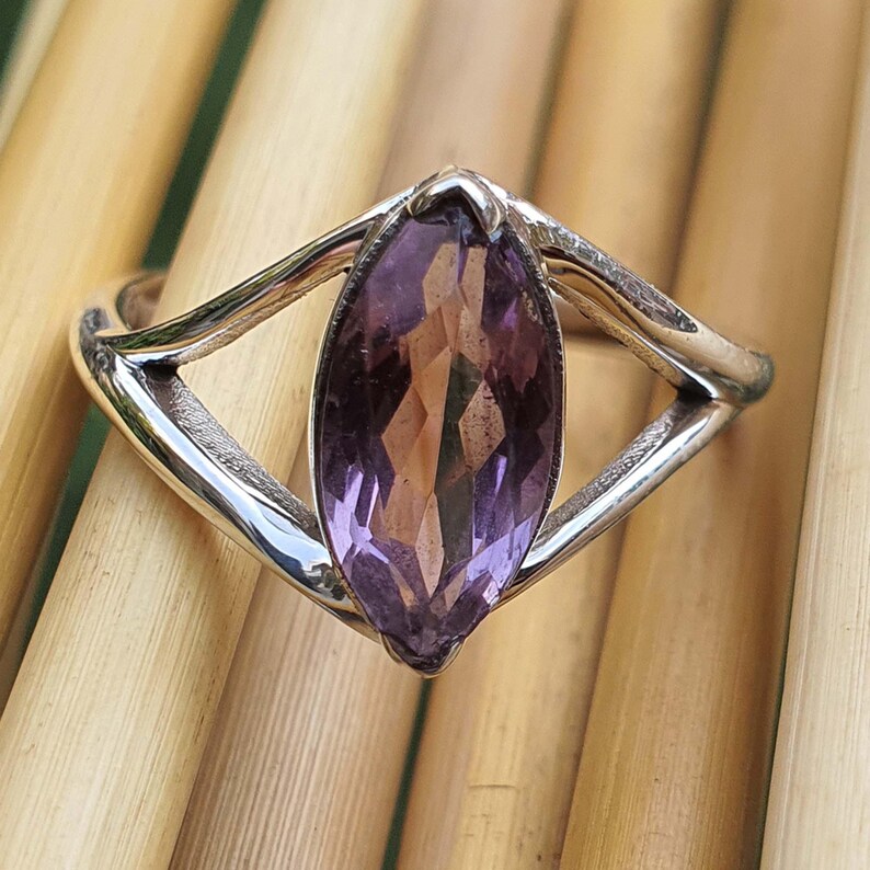 May include: A silver ring featuring a marquise-cut amethyst gemstone. The purple gemstone is set in a polished silver band with a unique, open design. The ring is displayed on a light brown surface.