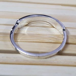 May include: A silver ring with a curved design. The ring is made of a shiny, reflective metal and has a smooth, polished surface. The ring is set against a light brown wooden background.