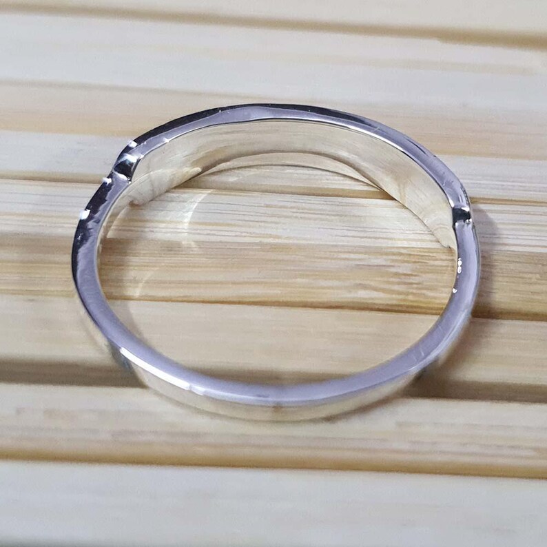 May include: A silver ring with a curved design. The ring is made of a shiny, reflective metal and has a smooth, polished surface. The ring is set against a light brown wooden background.