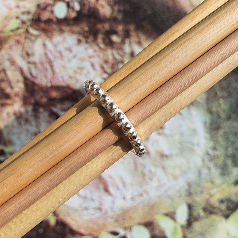 Op de afbeelding: Een zilveren ring met een kralenontwerp. De ring is gemaakt van een glanzend metaal en heeft kleine, gelijkmatig verdeelde kralen rond de band. De ring wordt getoond op verschillende houten deuvels.