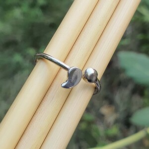 Pode incluir: Um anel aberto de tom prateado com duas extremidades distintas: uma em forma de pequena gota ou v&iacute;rgula lisa e a outra como uma esfera polida simples. A banda &eacute; fina e met&aacute;lica, concebida com um espa&ccedil;o ajust&aacute;vel entre as duas extremidades.
