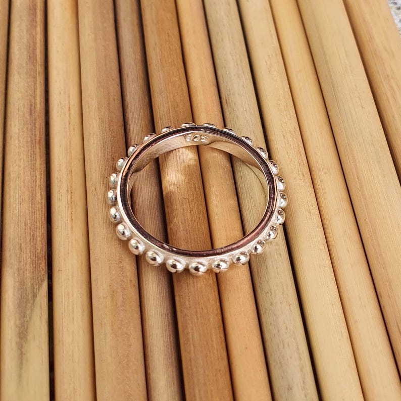 Op de afbeelding: Een zilveren ring met een kralenontwerp rond de band. De ring ligt op een lichtbruin houten oppervlak. De ring heeft een kleine inscriptie aan de binnenkant.