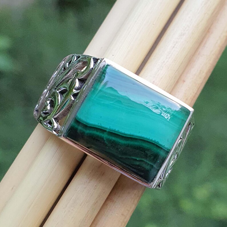 May include: A silver ring with a rectangular malachite stone. The stone is a vibrant green with darker green bands. The ring has an ornate, engraved design on the sides.