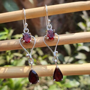Puede incluir: Un par de pendientes colgantes de plata con piedras preciosas rojas de forma ovalada y de l&aacute;grima. Los pendientes tienen un dise&ntilde;o en forma de coraz&oacute;n y est&aacute;n suspendidos de un gancho. Las piedras preciosas son de color rojo intenso.