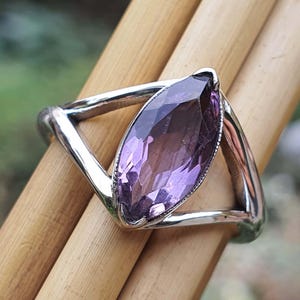 May include: A silver ring featuring a marquise-cut, faceted purple gemstone. The gemstone is set in a polished silver band with a unique, open design. The ring is displayed on a wooden surface, showcasing its elegant simplicity.