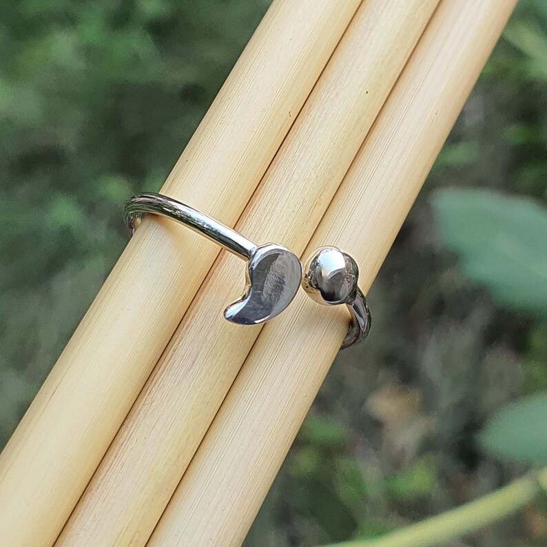 Pode incluir: Um anel aberto de tom prateado com duas extremidades distintas: uma em forma de pequena gota ou v&iacute;rgula lisa e a outra como uma esfera polida simples. A banda &eacute; fina e met&aacute;lica, concebida com um espa&ccedil;o ajust&aacute;vel entre as duas extremidades.