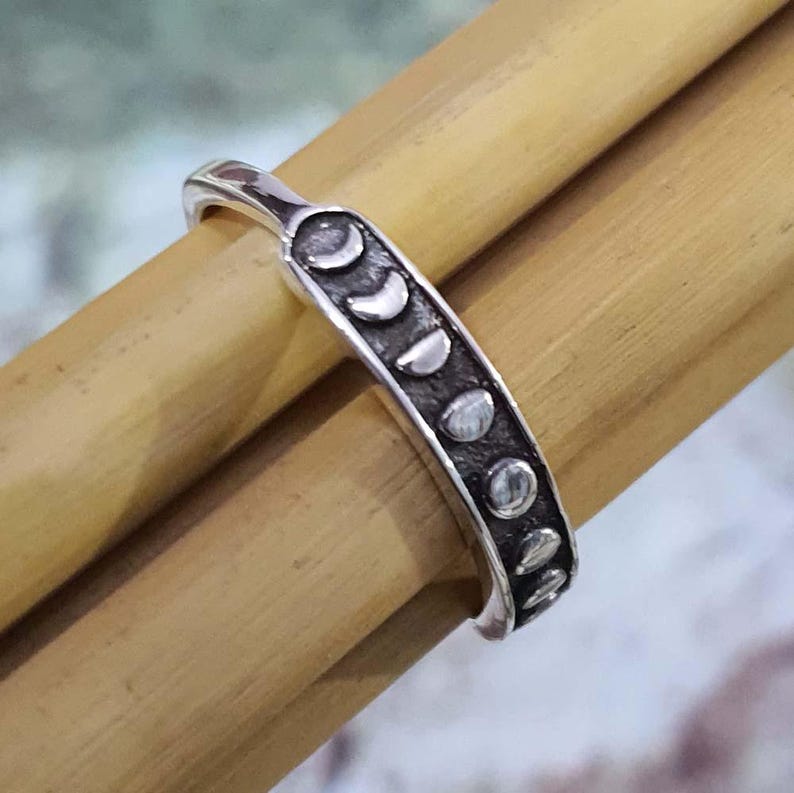 May include: A silver ring featuring a series of moon phase designs. The ring is a band style, with the moon phases arranged in a row along the top. The ring is shown on a light brown surface.