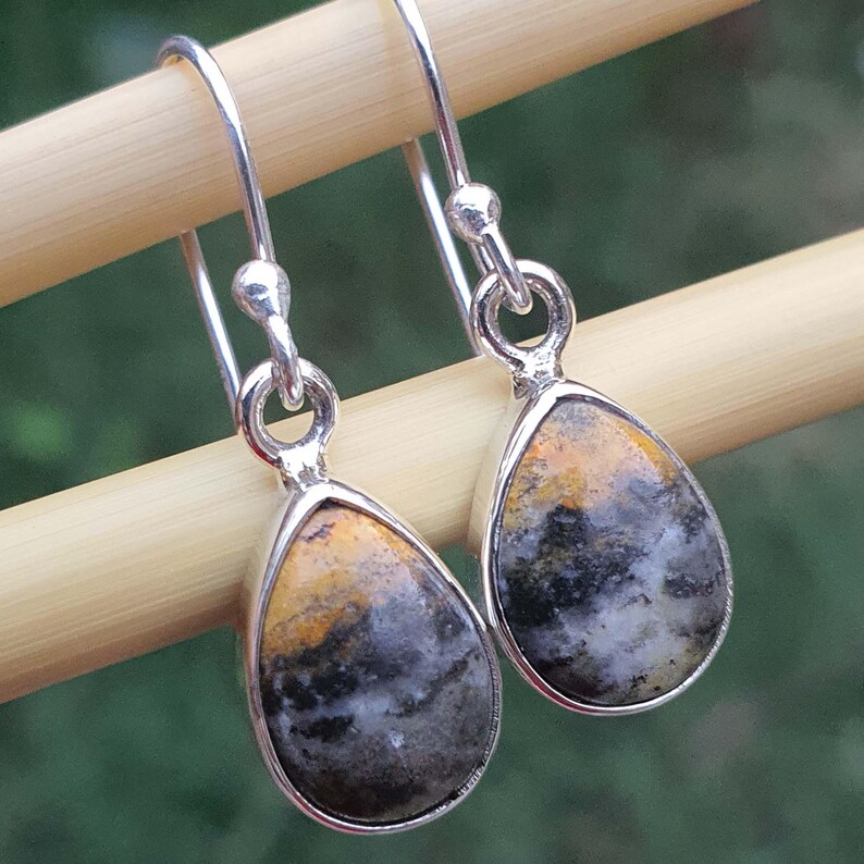 May include: A pair of silver drop earrings with teardrop-shaped stones. The stones feature a mix of yellow, gray, and black colors. The earrings are suspended from silver hooks and are displayed against a blurred green background.