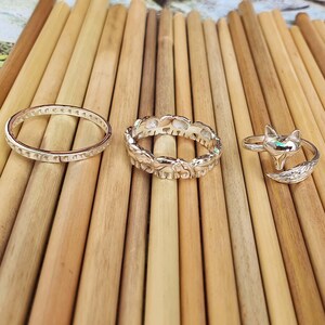 May include: Three silver rings displayed on a wooden surface. The rings include a simple band, a band with elephant designs, and a fox-shaped ring with a small green stone. The rings are made of shiny metal.