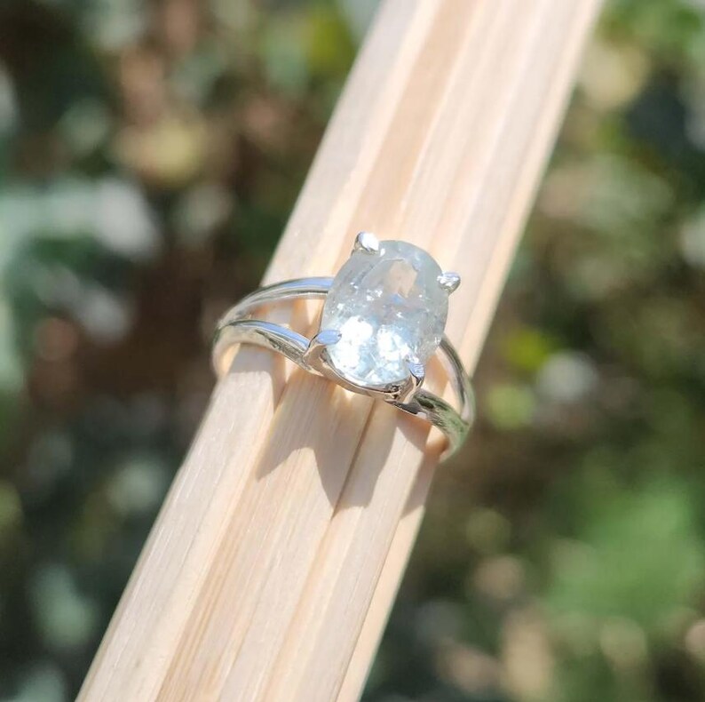 May include: A silver ring featuring a large, oval-cut, translucent light blue gemstone held in a four-prong setting. The band is split into two thin, polished silver segments that meet at the base of the stone, creating a sleek, open design.