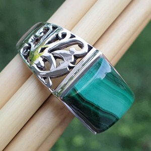 May include: A silver-toned ring with a rectangular, polished malachite stone. The ring features an openwork design with intricate patterns. The malachite stone displays bands of dark and light green. The ring is displayed on a light brown surface.