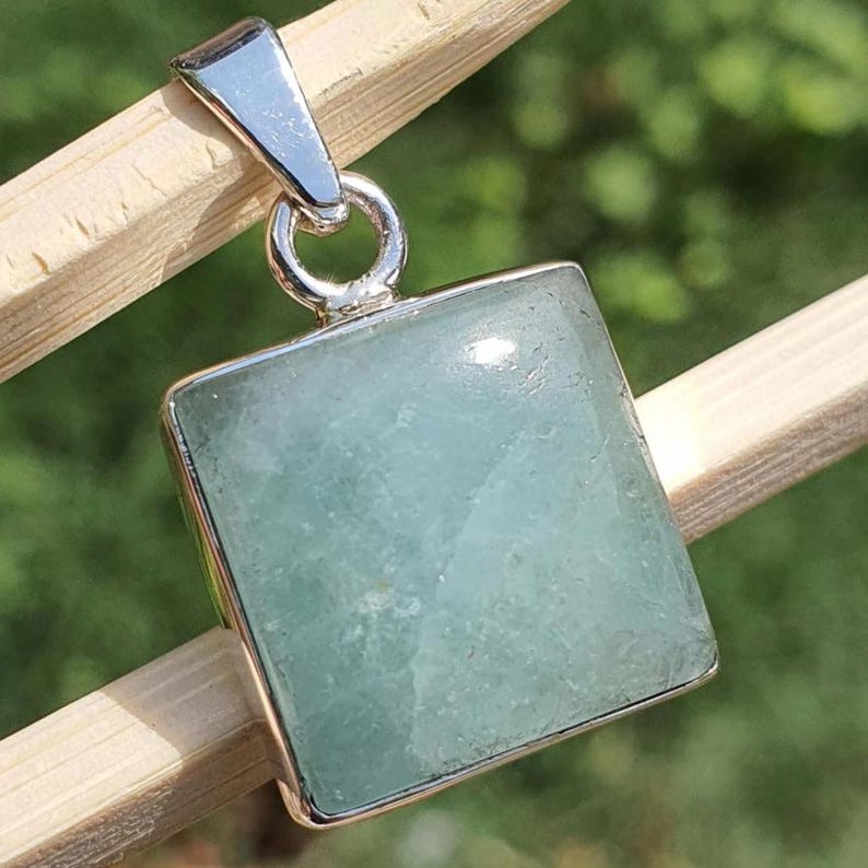 May include: A square-shaped pendant featuring a pale green, translucent gemstone set in a polished silver-toned metal frame. The pendant includes a silver-toned metal bail at the top, designed for attachment to a chain or cord for wear as jewelry.