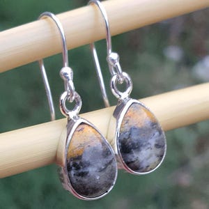 May include: A pair of silver drop earrings with teardrop-shaped stones. The stones feature a mix of colors, including black, gray, and yellow. The earrings are suspended from silver hooks.