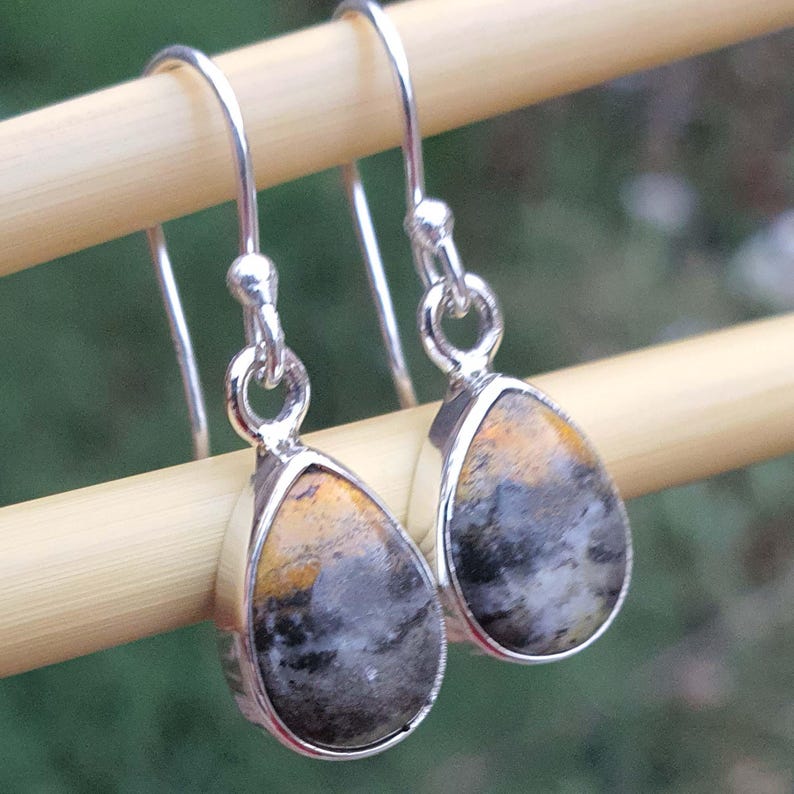 May include: A pair of silver drop earrings with teardrop-shaped stones. The stones feature a mix of colors, including black, gray, and yellow. The earrings are suspended from silver hooks.