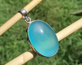 May include: A silver pendant featuring an oval, light blue gemstone. The gemstone is set in a gold-coloured bezel and has a smooth, polished surface. The pendant is suspended from a silver bail.