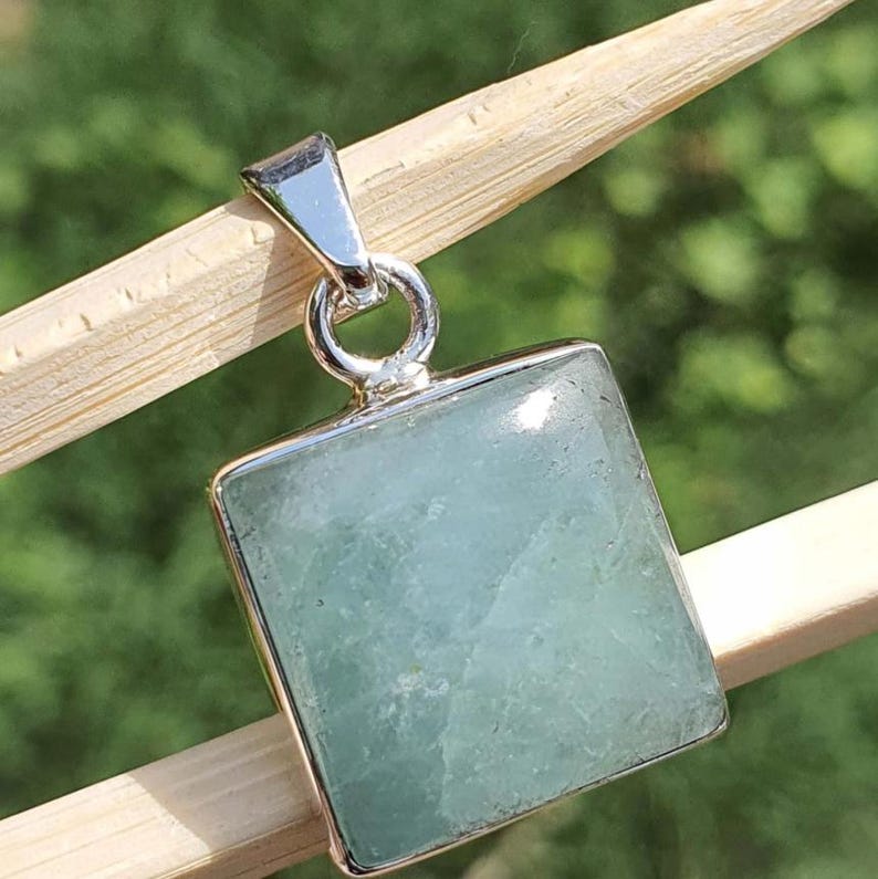 May include: A square-shaped pendant featuring a pale, translucent light green gemstone set in a polished silver-toned metal frame. The pendant includes a silver-toned metal bail at the top, designed for attachment to a necklace chain or cord.