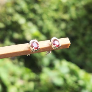 May include: Two stud earrings with oval, deep red gemstones set in silver metal. The earrings are presented on a light brown surface, with a blurred green backdrop. The setting suggests natural light.