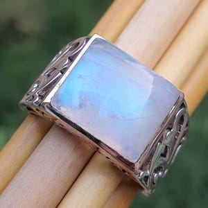 May include: A silver ring with a large, rectangular, iridescent moonstone. The ring has an ornate, engraved design on the band. The moonstone displays a soft, blue and white color, and is the focal point of the jewelry.