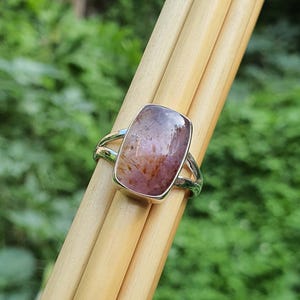 May include: A silver ring with a rectangular, purple and brown gemstone. The ring is set on a wooden surface.