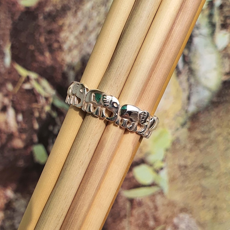 May include: A silver ring featuring a repeating elephant design. The ring has a polished finish and is adorned with small, green accents. The elephants are arranged in a continuous band, creating a unique and eye-catching piece of jewelry.