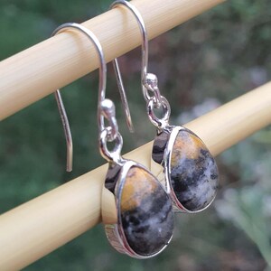 May include: Silver-tone drop earrings with teardrop-shaped stones. The stones feature a mix of yellow, black, and gray colors. The earrings are displayed on a light brown wooden dowel against a blurred green background.