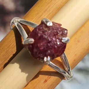 May include: A silver ring featuring a large, raw, deep red ruby gemstone. The ruby is held in place by four silver prongs. The ring's band splits into two delicate silver strands on each side, creating an open design.