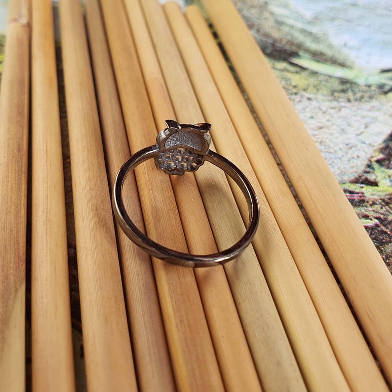 May include: A silver-colored ring with an owl-shaped design. The ring is set on a light brown surface. The owl's head is detailed with textured features.