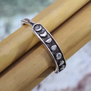 May include: A silver ring featuring a series of moon phase designs. The ring is a band style, with the moon phases arranged in a row along the top. The ring is shown on a light brown surface.