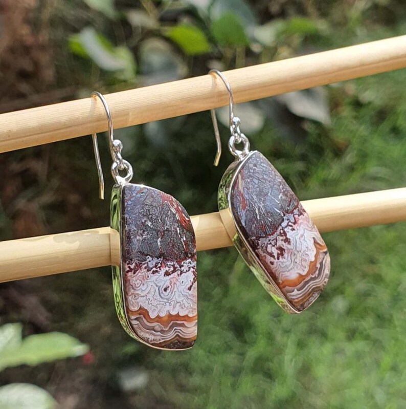 May include: A pair of dangle earrings featuring polished, irregular, rectangular-shaped stones with banded patterns in shades of dark brown, cream, and rust orange. The stones are set in silver-toned metal frames and hang from silver-toned wire hooks.