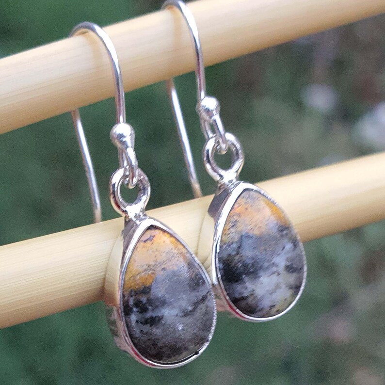 May include: A pair of silver drop earrings with teardrop-shaped stones. The stones feature a mix of colors, including black, gray, and yellow. The earrings are suspended from silver hooks.