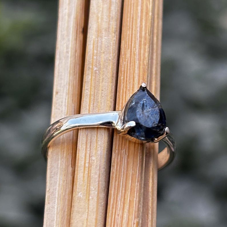 May include: A gold-toned ring featuring a pear-shaped, dark blue gemstone. The gemstone is set in a prong setting, and the band has a smooth, polished finish. The ring is displayed against a backdrop of wooden sticks.