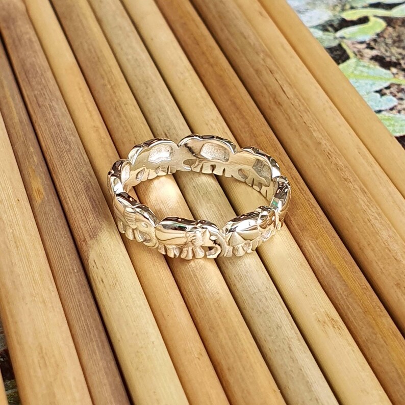 May include: A silver ring featuring a repeating pattern of elephant silhouettes. The ring is made of a shiny, metallic material and is set against a background of light brown wooden dowels.