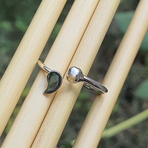 Pode incluir: Um anel aberto de tom prateado com duas extremidades distintas: um lado tem a forma de uma gota ou v&iacute;rgula lisa e polida, enquanto o outro lado termina numa pequena esfera arredondada e refletora. A superf&iacute;cie do metal &eacute; brilhante e polida.