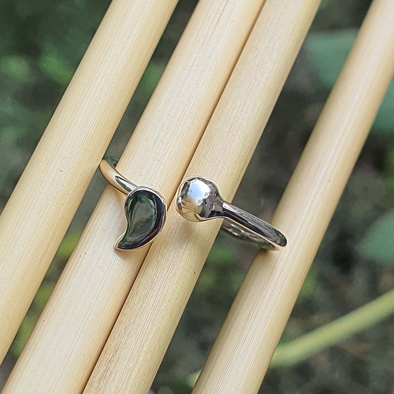 Pode incluir: Um anel aberto de tom prateado com duas extremidades distintas: um lado tem a forma de uma gota ou v&iacute;rgula lisa e polida, enquanto o outro lado termina numa pequena esfera arredondada e refletora. A superf&iacute;cie do metal &eacute; brilhante e polida.