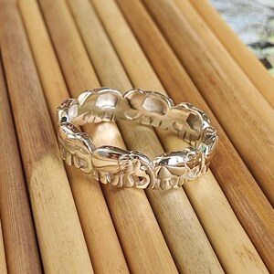 May include: A silver ring featuring a repeating pattern of elephant figures. The ring is made of a shiny, reflective metal and is set against a background of wooden dowels.