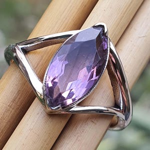 May include: A silver ring featuring a large, marquise-cut amethyst gemstone. The amethyst displays a gradient of purple hues, from deep violet to lighter shades. The ring's band has a geometric design, with a triangular shape on each side of the gemstone.