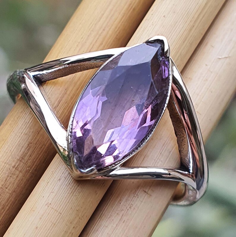 May include: A silver ring featuring a large, marquise-cut amethyst gemstone. The amethyst displays a gradient of purple hues, from deep violet to lighter shades. The ring's band has a geometric design, with a triangular shape on each side of the gemstone.