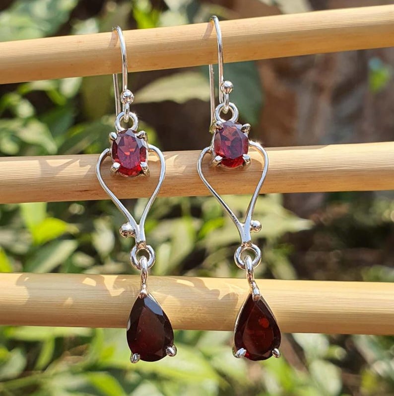 Puede incluir: Pendientes colgantes de plata con piedras rojas ovaladas y en forma de l&aacute;grima. Los pendientes tienen un dise&ntilde;o en forma de coraz&oacute;n y se muestran sobre un fondo verde borroso.