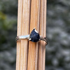 May include: A silver ring with a pear-shaped, dark blue gemstone. The ring is set on a wooden surface, showcasing the contrast between the shiny metal and the deep color of the stone. The ring's band is smooth and polished.