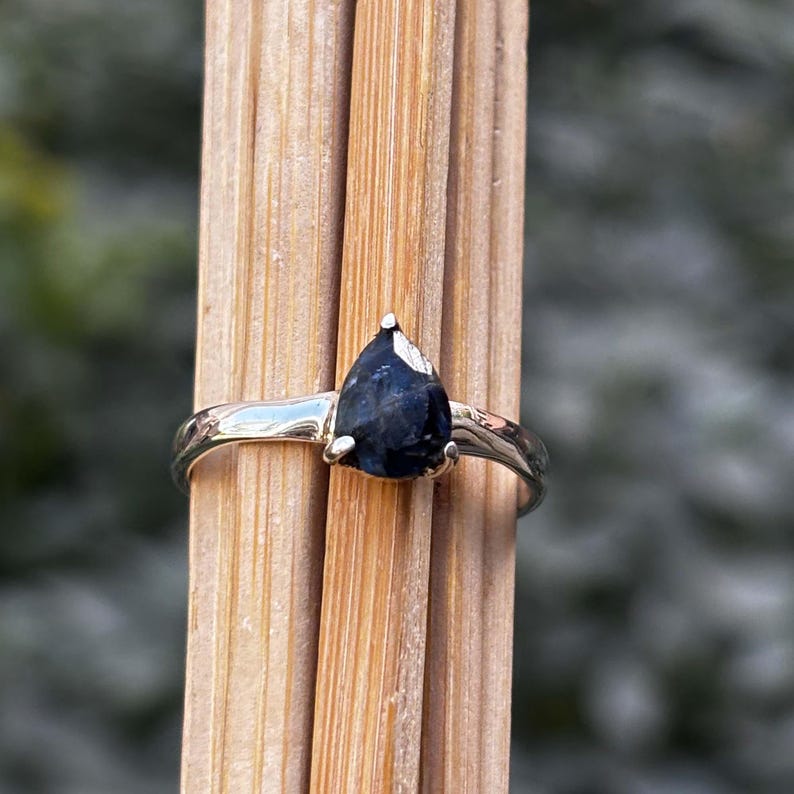May include: A silver ring with a pear-shaped, dark blue gemstone. The ring is set on a wooden surface, showcasing the contrast between the shiny metal and the deep color of the stone. The ring's band is smooth and polished.