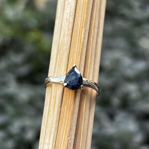 May include: A silver ring featuring a pear-shaped, dark blue gemstone. The ring's band has a simple, elegant design. The gemstone is securely set with small prongs, showcasing its polished surface. The ring is displayed against a backdrop of wooden sticks.