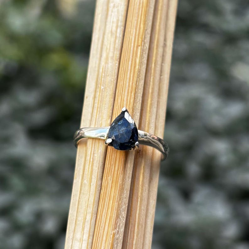 May include: A silver ring featuring a pear-shaped, dark blue gemstone. The ring's band has a simple, elegant design. The gemstone is securely set with small prongs, showcasing its polished surface. The ring is displayed against a backdrop of wooden sticks.