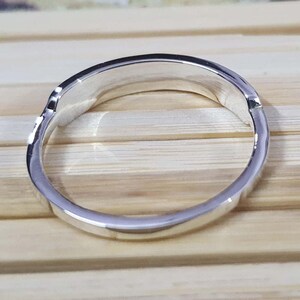 May include: A silver ring with a smooth, polished surface. The ring has a simple, elegant design, reflecting light. The ring is shown against a light brown wooden surface.