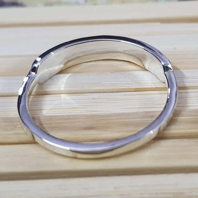 May include: A silver ring with a smooth, polished surface. The ring has a simple, elegant design, reflecting light. The ring is shown against a light brown wooden surface.