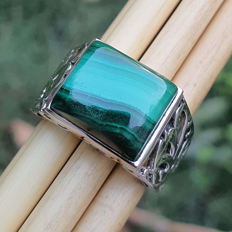 May include: A rectangular ring with a large, polished malachite stone in shades of green. The silver band features an ornate, openwork design. The ring is displayed on light-coloured wooden dowels against a blurred green background.